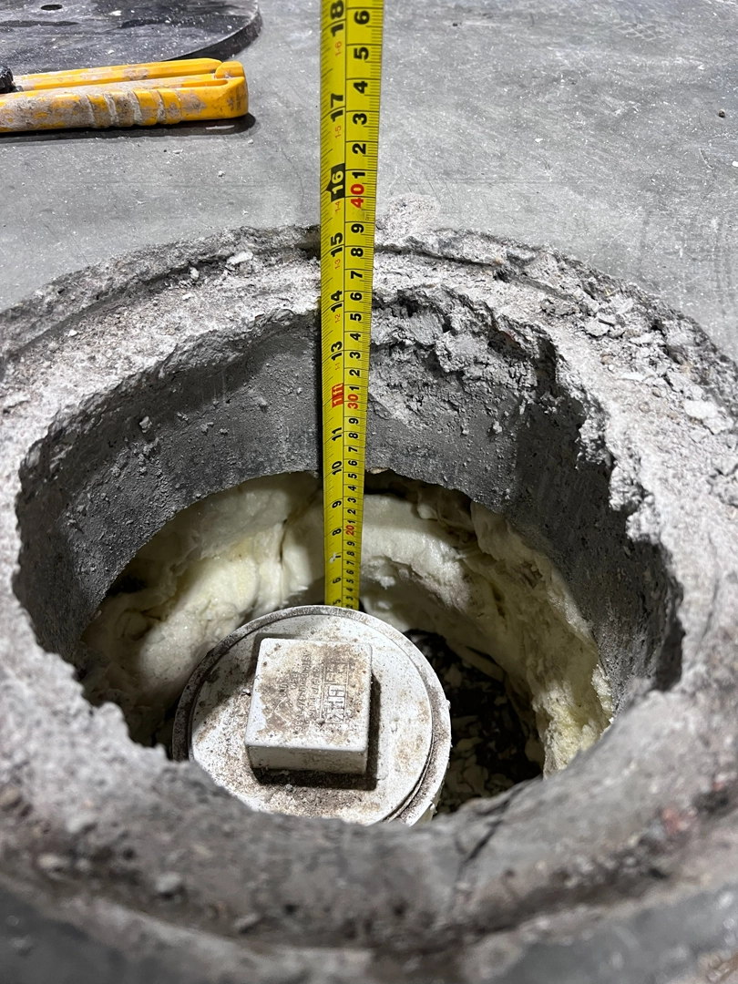 A close-up view of a yellow tape measure measuring the depth of a cored hole in a concrete floor down to a white pipe cap