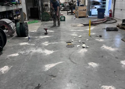 A worker uses a heavy-duty drill on the concrete floor of a large industrial workshop with various tools and materials around them