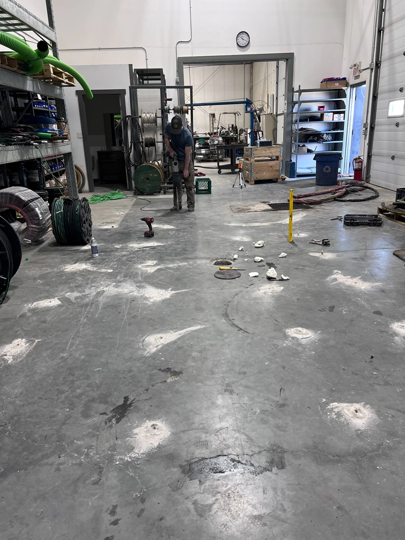 A worker uses a heavy-duty drill on the concrete floor of a large industrial workshop with various tools and materials around them
