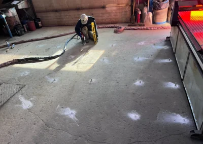A construction worker kneels on a concrete floor while installing anchor bolts with a power tool and a level