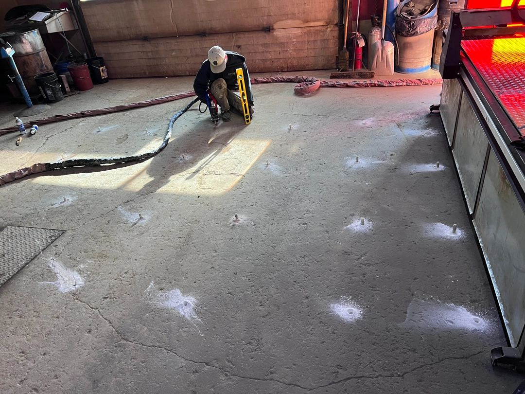 A construction worker kneels on a concrete floor while installing anchor bolts with a power tool and a level